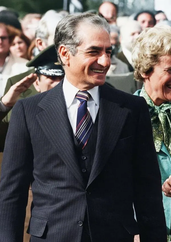 A smiling man in a pinstripe suit and tie stands among a crowd of people outdoors — a public domain photograph from The Shah of Iran archive documenting the Pahlavi era.