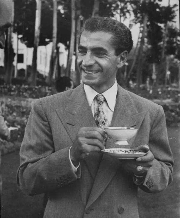 A smiling man in a suit and tie holds a teacup and saucer outdoors near trees — a public domain photograph from The Shah of Iran archive documenting the Pahlavi era.
