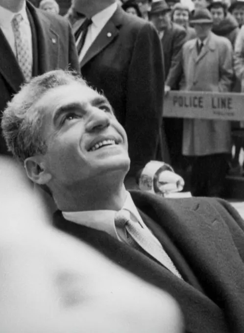 A smiling man in a suit looks upward while other men in formal attire stand behind him — a public domain photograph from The Shah of Iran archive documenting the Pahlavi era.