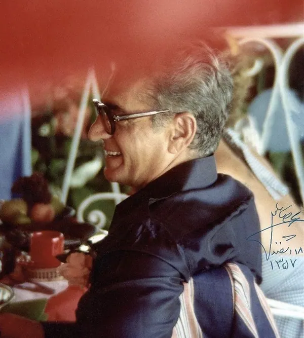 A smiling man with glasses and a dark jacket sits at a table outdoors — a public domain photograph from The Shah of Iran archive documenting the Pahlavi era.