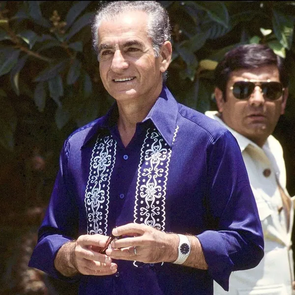 A smiling middle-aged man in a blue embroidered shirt stands before another man wearing sunglasses — a public domain photograph from The Shah of Iran archive documenting the Pahlavi era.