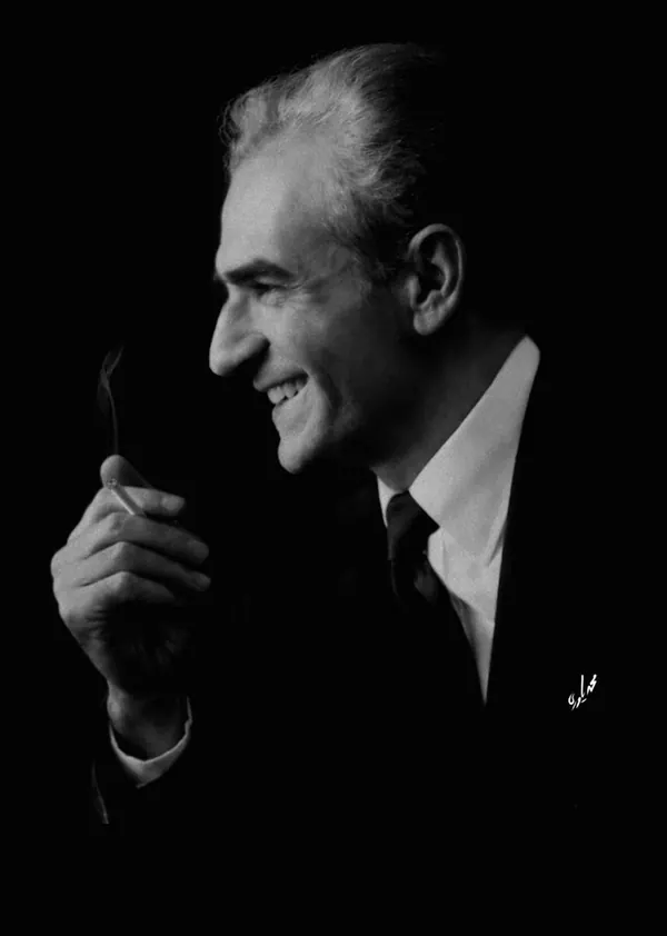 A smiling middle-aged man in a dark suit and tie holds a lit cigarette in his hand — a public domain photograph from The Shah of Iran archive documenting the Pahlavi era.
