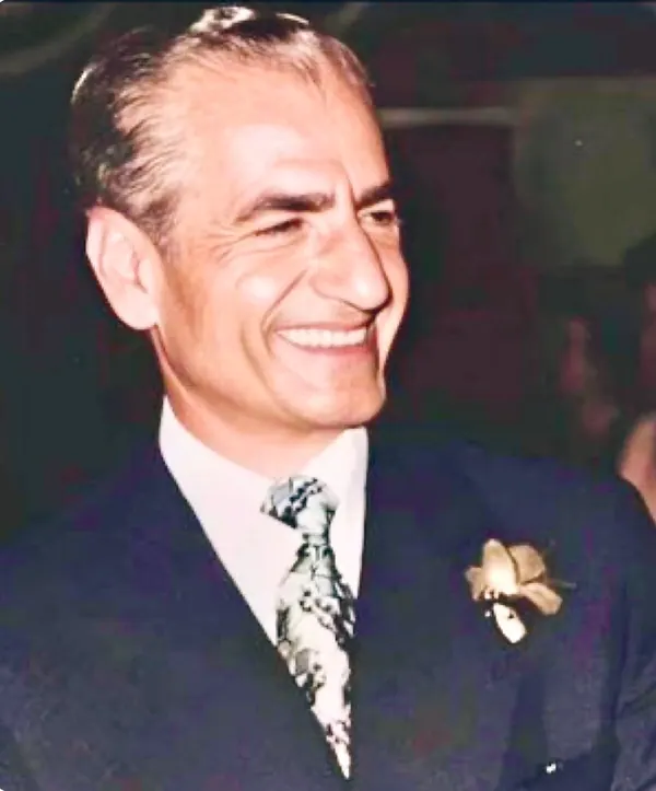 A smiling middle-aged man wears a dark suit, a patterned tie, and a flower on his lapel — a public domain photograph from The Shah of Iran archive documenting the Pahlavi era.