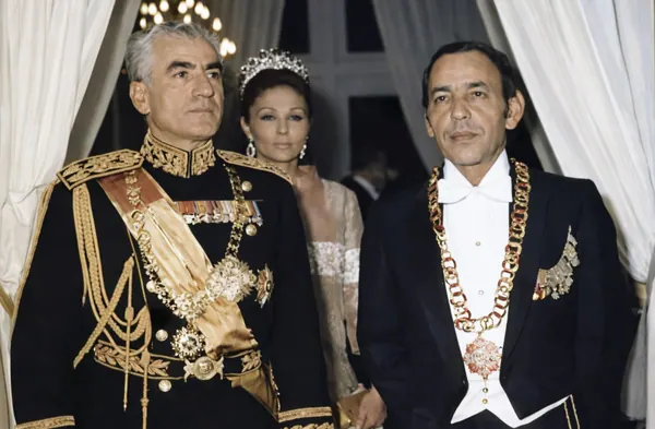 Three adults in formal attire and uniforms stand together indoors against a backdrop of white curtains — a public domain photograph from The Shah of Iran archive documenting the Pahlavi era.