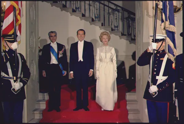 Three adults in formal attire stand on a red carpet between two uniformed guards in a grand interior — a public domain photograph from The Shah of Iran archive documenting the Pahlavi era.