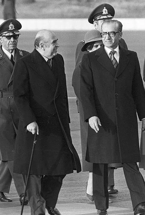 His Majesty, the Shah of Iran, and others in dark coats and uniforms walk outdoors, with one man holding a walking cane — a public domain photograph from The Shah of Iran archive documenting the Pahlavi era.