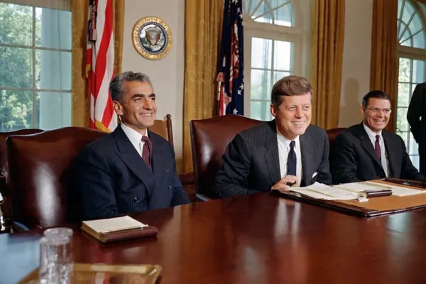 His Majesty, the Shah of Iran, and others in dark suits sit behind a large wooden table in a room with gold curtains — a public domain photograph from The Shah of Iran archive documenting the Pahlavi era.