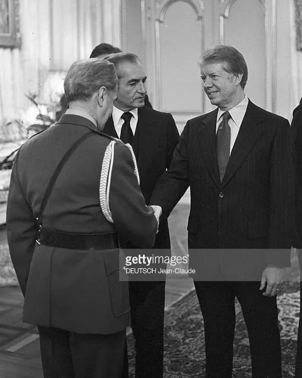 His Majesty, the Shah of Iran, and others in formal attire stand indoors, with two of them shaking hands in a medium shot — a public domain photograph from The Shah of Iran archive documenting the Pahlavi era.