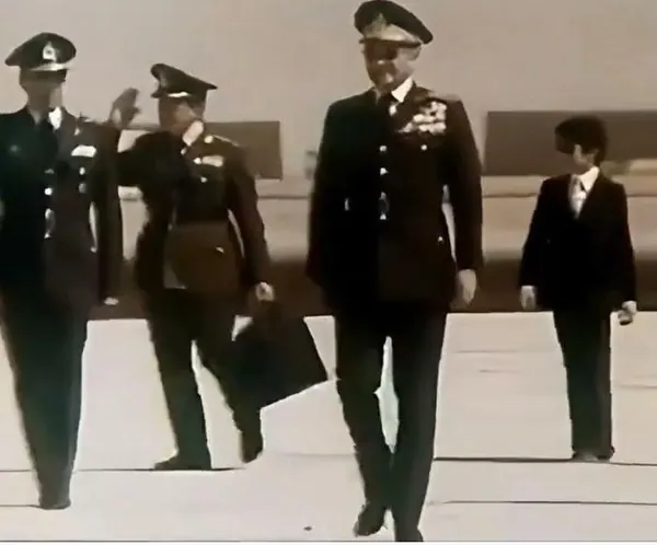 His Majesty, the Shah of Iran, and others in military uniforms and one man in a suit walk across a flat open area — a public domain photograph from The Shah of Iran archive documenting the Pahlavi era.