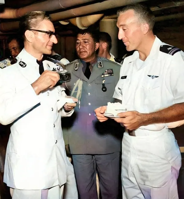 His Majesty, the Shah of Iran, and others in military uniforms stand together indoors, with two holding small plates of food — a public domain photograph from The Shah of Iran archive documenting the Pahlavi era.