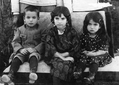 Three young children in patterned and structured clothing sit side by side on a stone ledge — a public domain photograph from The Shah of Iran archive documenting the Pahlavi era.