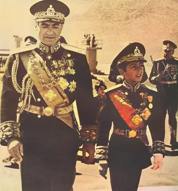 Two males in ornate dark military uniforms with gold braids and sashes stand outdoors in a sandy area — a public domain photograph from The Shah of Iran archive documenting the Pahlavi era.
