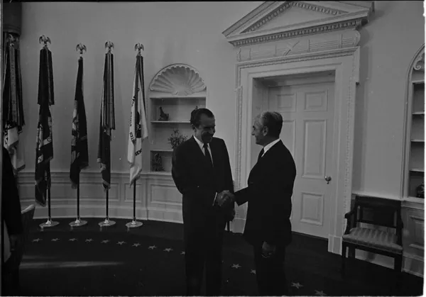 His Majesty, the Shah of Iran, and others in dark suits shake hands in a room with flags and a white door — a public domain photograph from The Shah of Iran archive documenting the Pahlavi era.