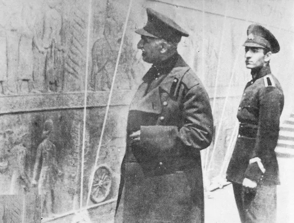 His Majesty, the Shah of Iran, and others in military uniforms and caps stand before a wall with carved relief figures — a public domain photograph from The Shah of Iran archive documenting the Pahlavi era.