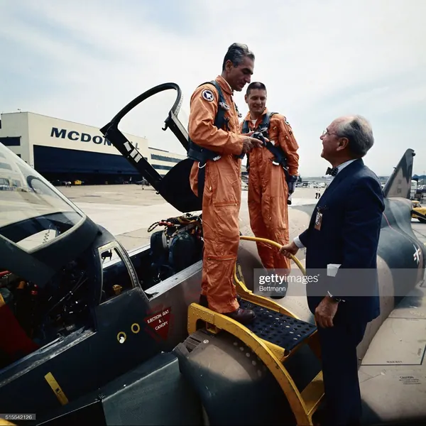 His Majesty, the Shah of Iran, and others in orange flight suits and one man in a suit stand beside a jet aircraft — a public domain photograph from The Shah of Iran archive documenting the Pahlavi era.