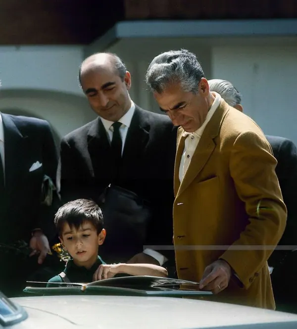 His Majesty, the Shah of Iran, and others in suits and a young boy look down at an open book on a car hood — a public domain photograph from The Shah of Iran archive documenting the Pahlavi era.