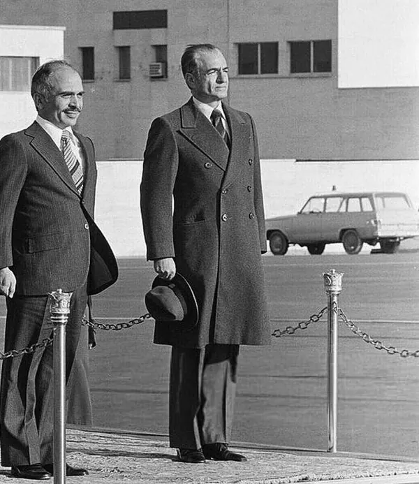 His Majesty, the Shah of Iran, and others in suits and overcoats stand behind a chain barrier outdoors near a parked car — a public domain photograph from The Shah of Iran archive documenting the Pahlavi era.
