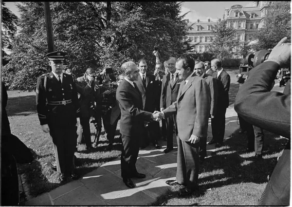 His Majesty, the Shah of Iran, and others in suits shake hands outdoors while surrounded by other men and a uniformed officer — a public domain photograph from The Shah of Iran archive documenting the Pahlavi era.
