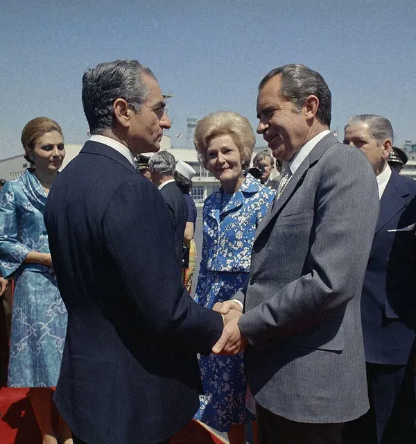 His Majesty, the Shah of Iran, and others in suits shake hands while Farah Pahlavi, Empress of Iran, in a blue floral dress looks on outdoors — a public domain photograph from The Shah of Iran archive documenting the Pahlavi era.