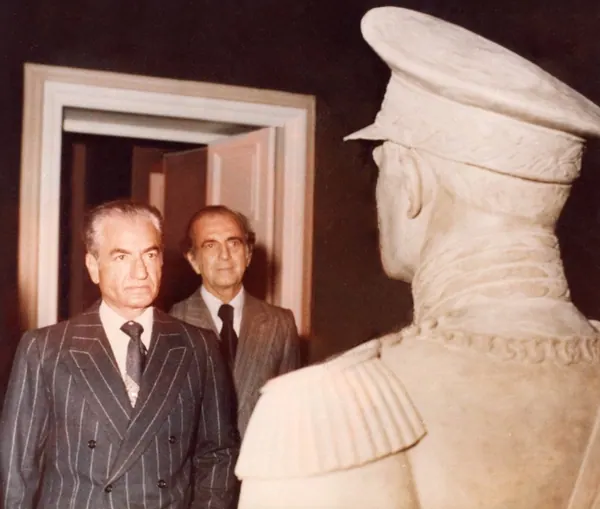 His Majesty, the Shah of Iran, and others in suits stand beside a large stone bust of His Majesty in a military cap — a public domain photograph from The Shah of Iran archive documenting the Pahlavi era.
