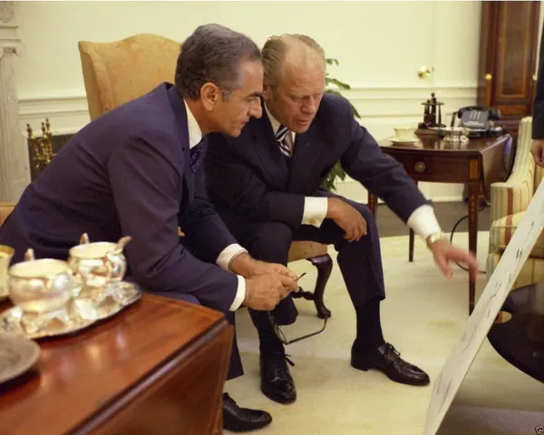 Two middle-aged men in dark suits lean forward to look at a large white board in a room — a public domain photograph from The Shah of Iran archive documenting the Pahlavi era.