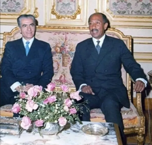 Two middle-aged men in dark suits sit on an ornate sofa behind a bouquet of pink roses — a public domain photograph from The Shah of Iran archive documenting the Pahlavi era.