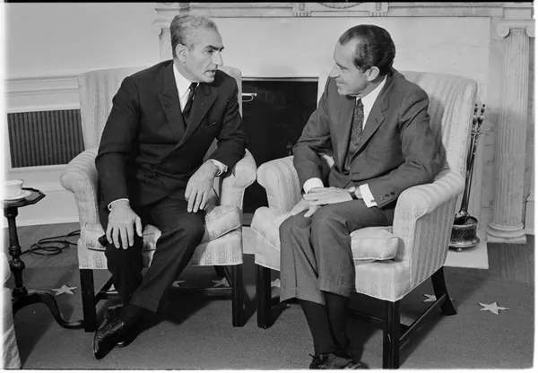 Two middle-aged men in dark suits sit in armchairs and talk in a room with a fireplace — a public domain photograph from The Shah of Iran archive documenting the Pahlavi era.