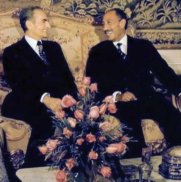 Two middle-aged men in dark suits sit on an ornate sofa behind a bouquet of pink roses — a public domain photograph from The Shah of Iran archive documenting the Pahlavi era.
