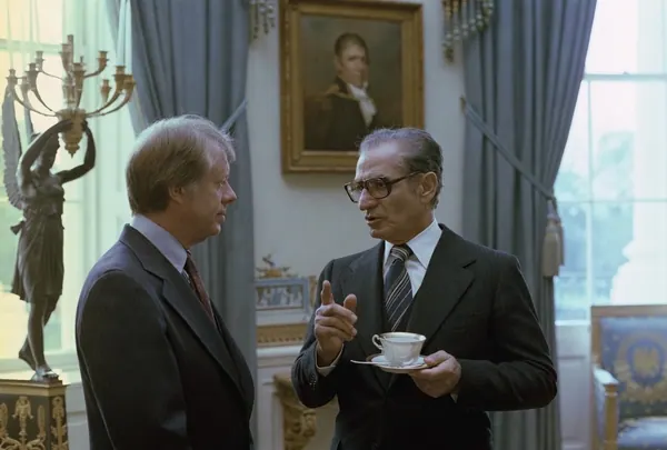 Two middle-aged men in dark suits talk in a room with a painting and a statue — a public domain photograph from The Shah of Iran archive documenting the Pahlavi era.