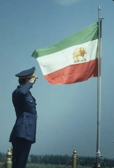A uniformed person in a blue cap and jacket salutes a tricolor flag flying on a pole — a public domain photograph from The Shah of Iran archive documenting the Pahlavi era.