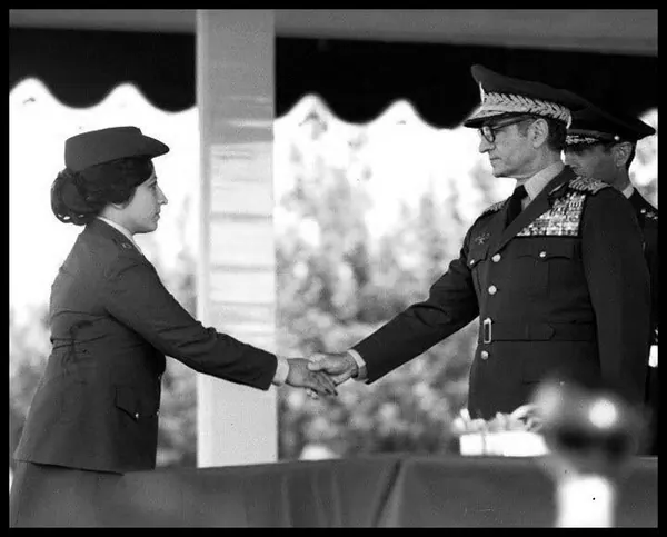 Farah Pahlavi, Empress of Iran, and His Majesty in military uniforms shake hands outdoors under a covered structure — a public domain photograph from The Shah of Iran archive documenting the Pahlavi era.