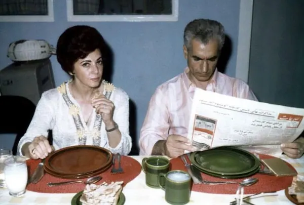 Farah Pahlavi, Empress of Iran, and His Majesty sit at a dining table with plates and a newspaper — a public domain photograph from The Shah of Iran archive documenting the Pahlavi era.