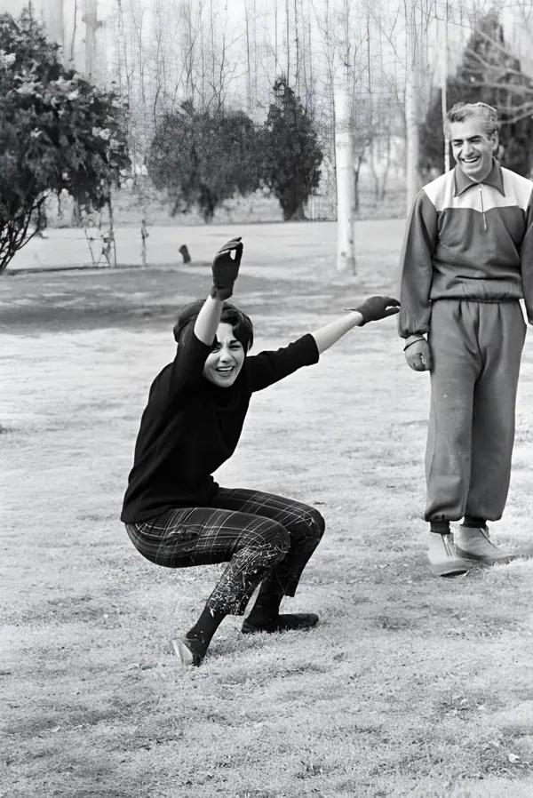Farah Pahlavi, Empress of Iran, in a black top and plaid pants squats while His Majesty in a tracksuit watches nearby — a public domain photograph from The Shah of Iran archive documenting the Pahlavi era.