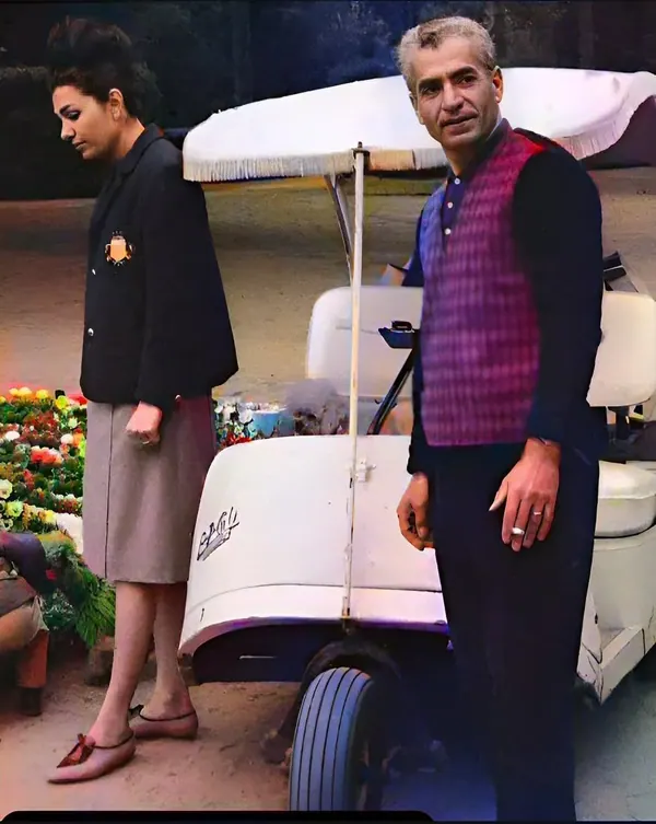 Farah Pahlavi, Empress of Iran, in a dark blazer and skirt stands beside His Majesty wearing a purple patterned vest — a public domain photograph from The Shah of Iran archive documenting the Pahlavi era.