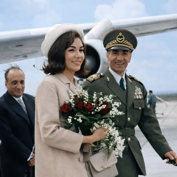 Farah Pahlavi, Empress of Iran, in a light coat and hat holds flowers beside His Majesty in a military uniform near a plane — a public domain photograph from The Shah of Iran archive documenting the Pahlavi era.
