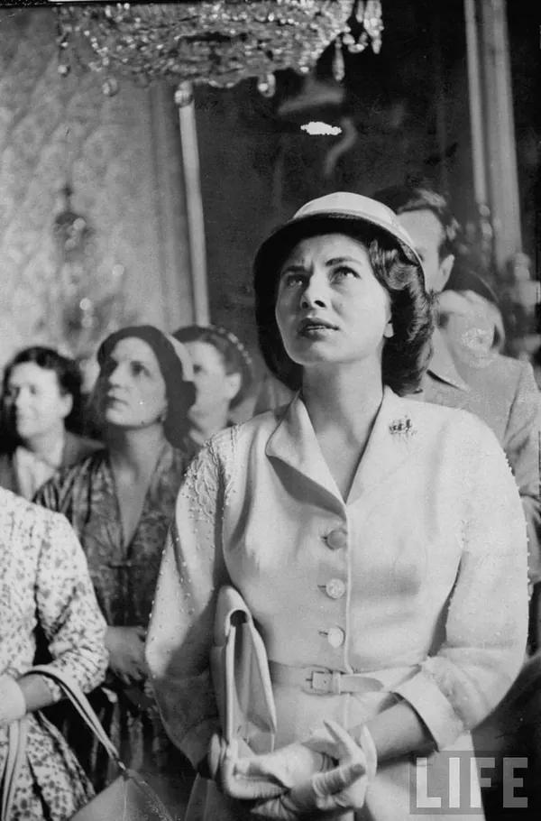 Farah Pahlavi, Empress of Iran, in a light-colored suit and hat looks upward while standing among other people indoors — a public domain photograph from The Shah of Iran archive documenting the Pahlavi era.