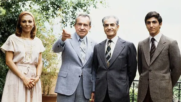 Farah Pahlavi, Empress of Iran, in a light dress and three men in suits stand together outdoors near green foliage — a public domain photograph from The Shah of Iran archive documenting the Pahlavi era.