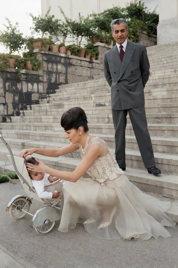 Farah Pahlavi, Empress of Iran, in a light dress tends to a child in a stroller while His Majesty stands behind — a public domain photograph from The Shah of Iran archive documenting the Pahlavi era.