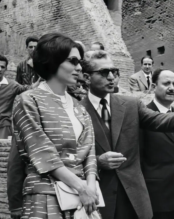 Farah Pahlavi, Empress of Iran, in a patterned suit and sunglasses stands beside His Majesty in a suit and glasses — a public domain photograph from The Shah of Iran archive documenting the Pahlavi era.