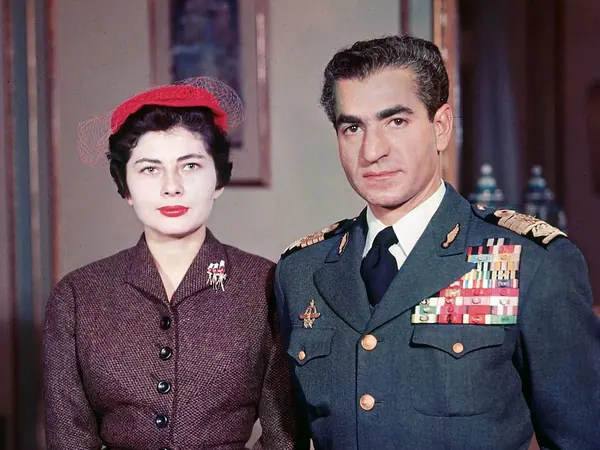 Farah Pahlavi, Empress of Iran, in a red hat and brown jacket stands beside His Majesty in a decorated military uniform — a public domain photograph from The Shah of Iran archive documenting the Pahlavi era.