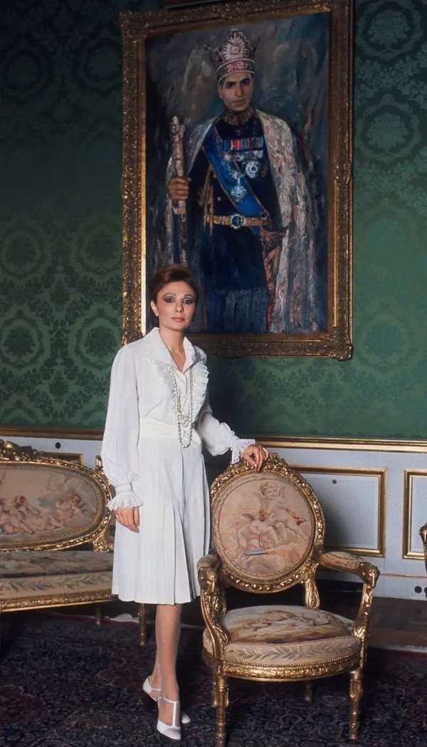 Farah Pahlavi, Empress of Iran, in a white dress stands beside an ornate chair beneath a large framed portrait of His Majesty — a public domain photograph from The Shah of Iran archive documenting the Pahlavi era.