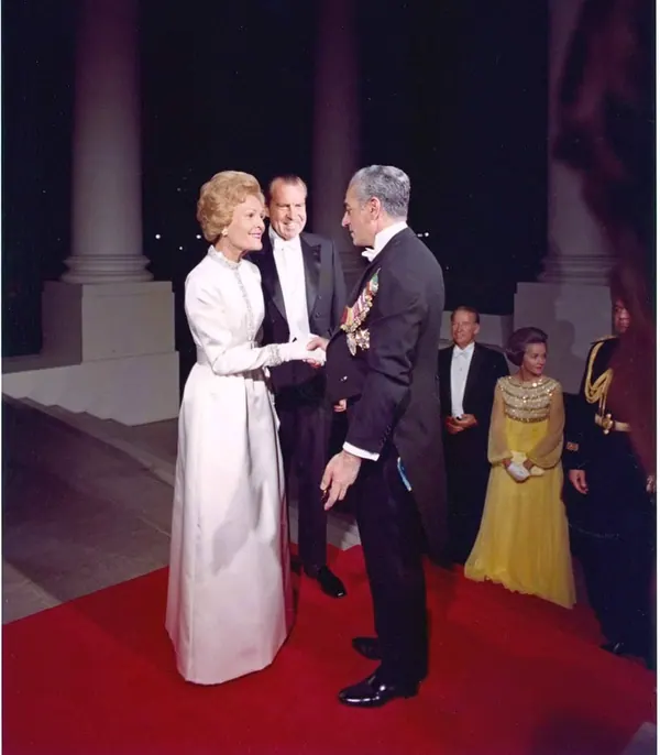 Farah Pahlavi, Empress of Iran, in a white gown shakes hands with His Majesty in a formal tuxedo on a red carpet — a public domain photograph from The Shah of Iran archive documenting the Pahlavi era.