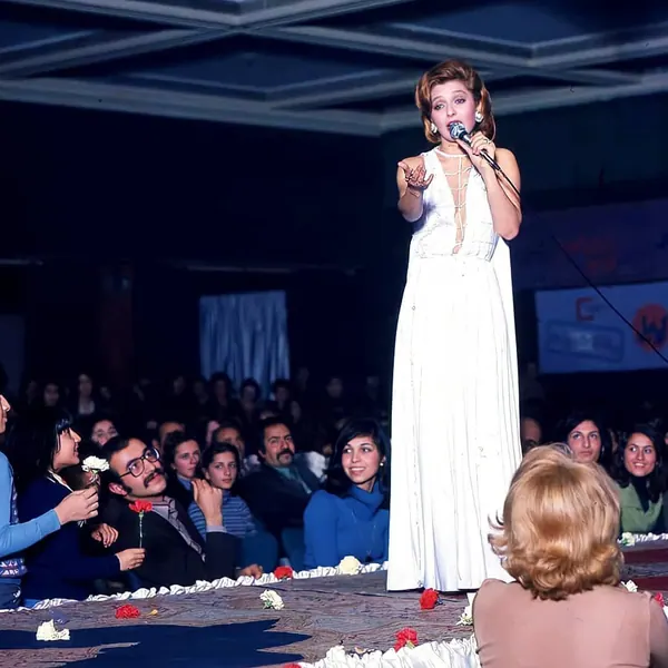 Farah Pahlavi, Empress of Iran, in a white gown sings into a microphone before a seated audience in a hall — a public domain photograph from The Shah of Iran archive documenting the Pahlavi era.