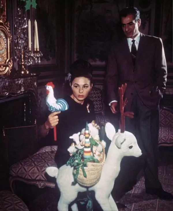 Farah Pahlavi, Empress of Iran, in black holds a rooster toy beside a white deer figure while His Majesty stands behind her — a public domain photograph from The Shah of Iran archive documenting the Pahlavi era.