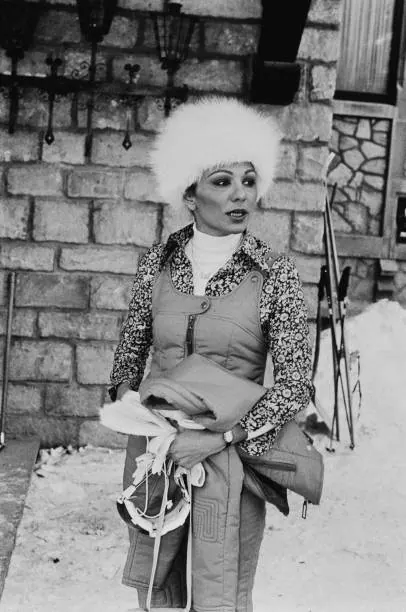 Farah Pahlavi, Empress of Iran, wearing a white fur hat and winter clothing stands before a stone wall with skis nearby — a public domain photograph from The Shah of Iran archive documenting the Pahlavi era.