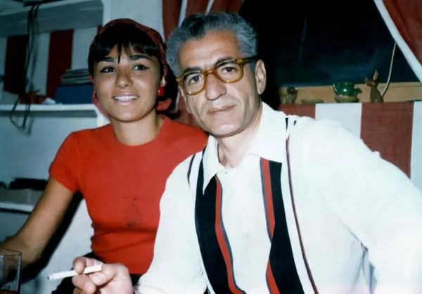 Farah Pahlavi, Empress of Iran, in a red shirt and His Majesty in glasses pose together indoors — a public domain photograph from The Shah of Iran archive documenting the Pahlavi era.